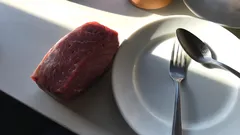 Syrový kus hovězího masa vedle talíře s vidličkou a lžící na bílém stole.